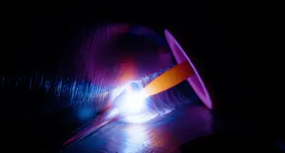 Schweißlichtbogen-Makro: glühende Elektrode und Schweißnaht in Blau- und Violetttönen auf dunklem Stahl
