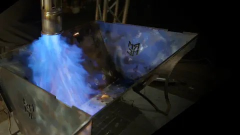 Blaue Flamme schlägt aus dem Anzündkamin in die Wanne des Schlitzers, das ME-Monogramm in der Seitenwange glüht im Feuerschein